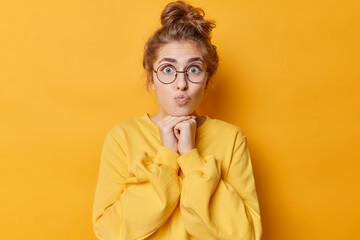 Portrait of surprised lovely woman keeps lips rounded hands under chin has dark hair gathered in bun wears round spectacles and jumper isolated over yellow background. Human face expressions