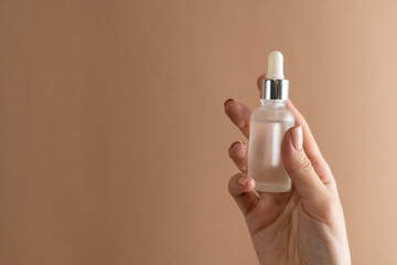 Graceful hand of a girl with thin fingers and a trendy pastel beige nude manicure on a brown background holds a face oil serum