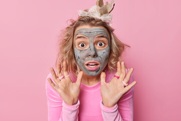 Scared emotional young woman raises palms in defensive gesture applies clay mask for skin treatment stands fearful wears crown annd jumper isolated over pink background. Omg what horrible scene