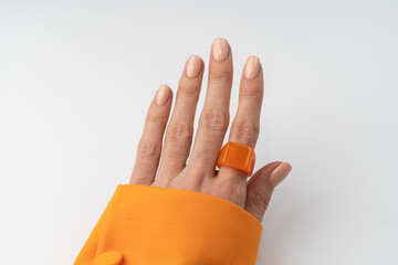 Close shot of a thin graceful hand of a girl with a nude pastel spring trendy manicure and a big ring on her finger in an orange jacket on a white background