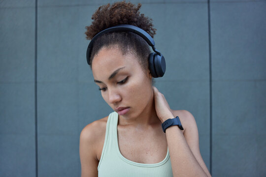 Tired Sporty Sweaty Woman Keeps Hand On Neck Feels Exhausted After Sport Training Dressed In T Shirt Looks Down Listens Music Via Headphones Poses Against Grey Wall Wears Wristwatch. Physical Activity