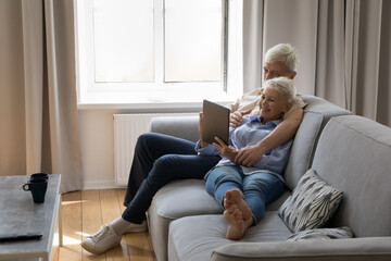Happy senior married couple resting on comfortable sofa together, using tablet computer, shopping on Internet, smiling, laughing, hugging, enjoying leisure time, digital gadget, making video call