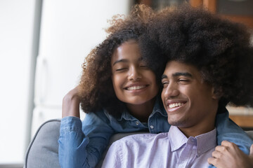 Happy young teen Black couple enjoying dating at home, embracing, talking, laughing. Cute teenage African girlfriend giving hug, cuddling fuzzy haired boyfriend with love, tenderness