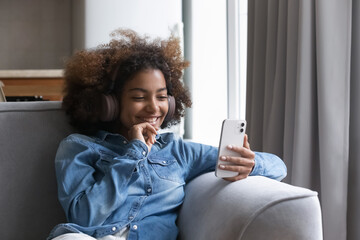 Cheerful pretty gen Z African teen girl listening to popular music, using smartphone, big...