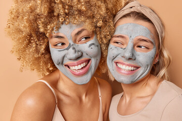 Close up portrait of two cheerful young women apply mud mask smile broadly show white teeth dressed in casual clothes isolated over brown background. Beauty procedures and skin care concept.