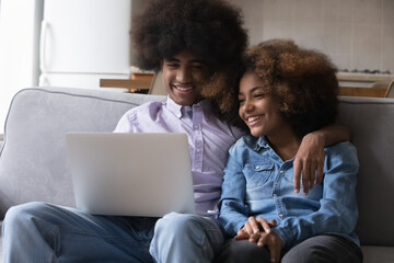 Happy teen dating Black couple enjoying leisure time at home, resting on couch, watching funny movie, sitcom, series on laptop online, using internet application for media files download
