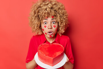 Impressed shocked young woman baked delicious heart shaped cake for husband on Vaentines Day looks with widely opened mouth and eyes at camera prepares for holiday isolated over red background