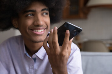 Fototapeta na wymiar Happy teenage young African American guy talking on cell on speaker, smiling, looking away. Smartphone user recording audio message, giving commands to virtual assistant. Hand with cellphone close up