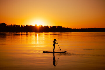 Rowing on sup board woman with oar in hands with her reflection on water on calm lake with soothing sunset and trees in background. Active lifestyle.