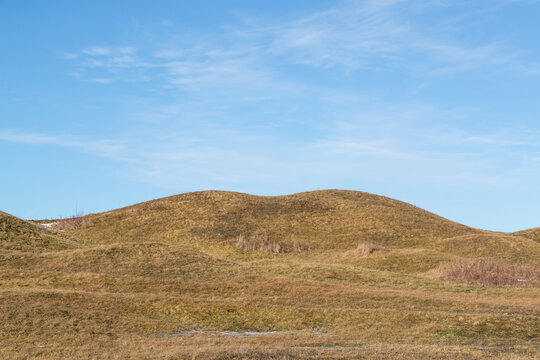 Burial mound covered with grass in rolling hill culture landscape. Patches of snow on the ground and clear sky with light clouds