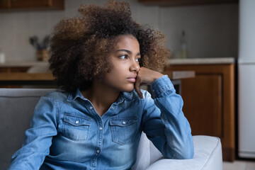 Frustrated pensive teenage African girl sitting on couch at home, looking away, thinking over...