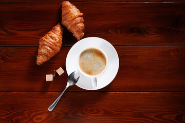 Croissants with a cup of coffee and two lumps of brown sugar lie on a wooden table. Focusing on coffee. View from above. Place for your text.