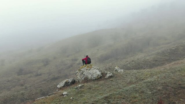 A man enjoys life sitting on a rock in the mountains and watching the fog in the mountains. Aerial Shot.