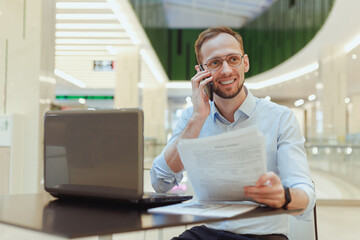 Smiling businessman man talking on the phone while working with documents on a laptop in a public place