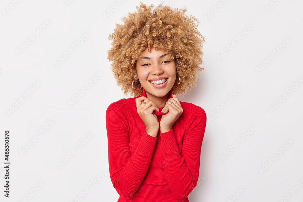 Wall mural Portrait of cheerful young woman with curly bushy hair keeps hands on collar of red jumper being in good mood feels very happy isolated over white background. Human emotions and sincere feelings - Wall murals