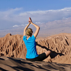 Woman doing relaxing yoga on dune in windy Atacama desert mountain landscape with impressive view flying hair