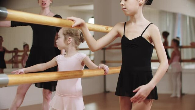 Little modest ballerina girls training in the studio near the stand with their female trainer