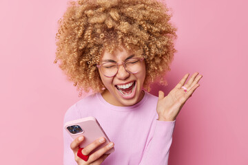 Photo of emotional woman with blonde curly hair exclaims loudly keeps palm raised uses mobile phone...
