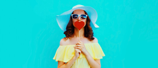 Fototapeta na wymiar Portrait of beautiful young woman blowing her lips with lipstick with red sweet heart shaped lollipop wearing a summer straw hat on blue background