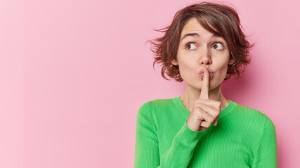 Beautiful young woman with bob hairstyle makes hush gesture shows silence sign wears green jumper...
