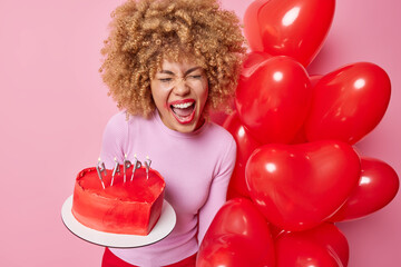 Emotional displeased curly woman exclaims loudly feels mad or depressed celebrates Valentines Day alone holds heart shaped cake and balloons hates this holiday poses against pink background.