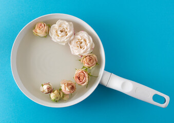 A creative spring summer concept made of a white frying pan with water and bright bush rose flowers on water. blue background.