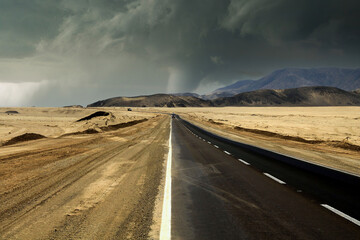 View on empty asphalt highway through arid dry landscape with dark storm clouds and tornado -...