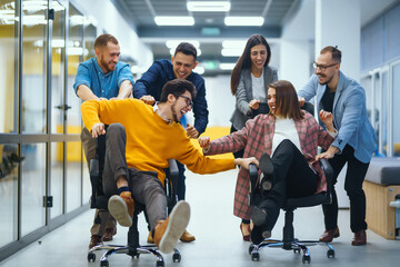 Fototapeta Young cheerful business people  having fun while racing on office chairs and smiling. Friendly work team enjoying fun work break activities. obraz