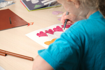 preschooler girl in a blue t-shirt makes an applique sitting at the table, hearts cut out of colored bougue for a card