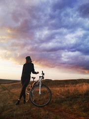 A beautiful tall girl stands with a bicycle against the background of a summer evening landscape. The concept of nature, freedom, sport, love of life.