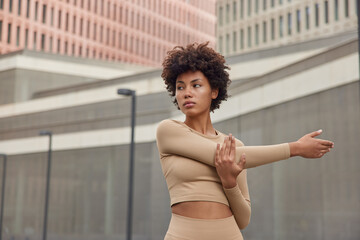 Waist up shot of active slim curly haired woman dressed in beige tracksuit stretches arms being in...