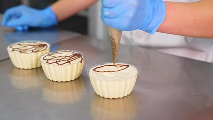 White creamy desserts being decorated with caramel. Gloved hand squeezes liquid caramel from pastry bag on the cups with sweets. Close up.