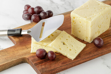 Cutting a piece of aromatic cheese with special slicer and red grapes  on wooden board on marble table in the kitchen. Light background with copy space