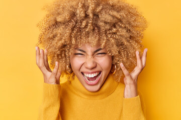 Fototapeta Headshot of emotional curly haired woman exclaims loudly keeps hands raised near head has mouth opened wears casual jumper isolated over vivid yellow background shouts from joy. Emotions concept obraz