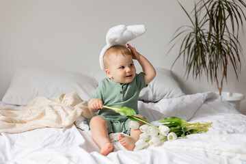 baby with Easter bunny ears in a green cotton bodysuit is sitting in bed with white tulips in his hands. products for children and holidays. space for text. High quality photo