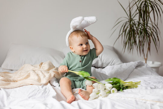 baby with Easter bunny ears in a green cotton bodysuit is sitting in bed with white tulips in his hands. products for children and holidays. space for text. High quality photo