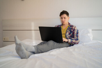 young caucasian man sits at home on the bed in the morning in casual clothes and visits internet sites using his laptop.