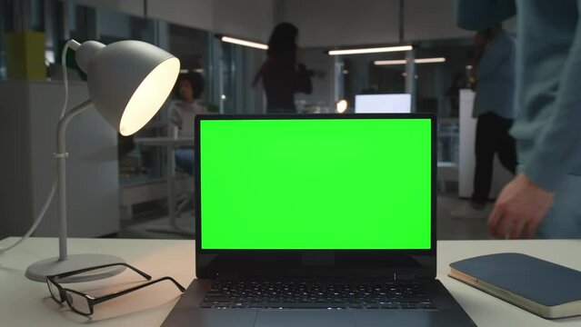 Laptop with green screen on desk in office with employees working on background