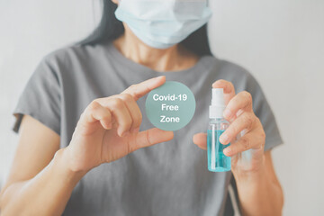 blue circle paper with COVID-19 Free Zone text and alcohol spray bottle in senior woman's hands with blurred face wearing medical mask for population, social or herd immunity concept
