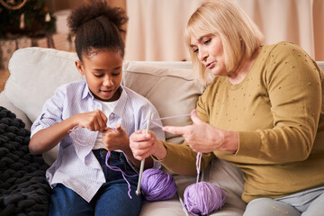 Senior woman feeling concentrated while knitting at the sofa with her multiracial child