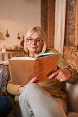 Senior woman holding book and feeling concentrated while reading at the sofa