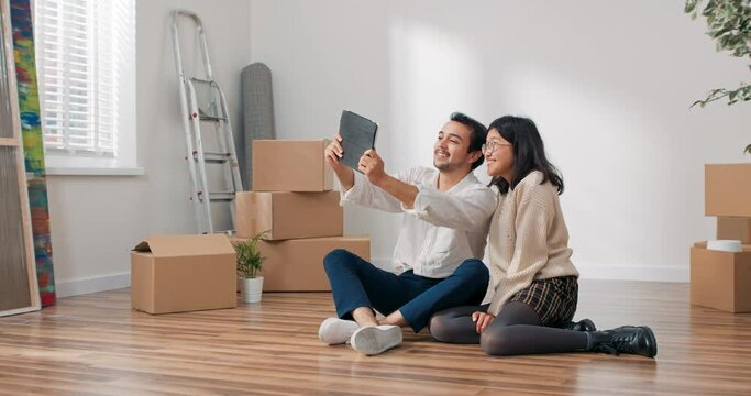 Lovers are sitting on floor in newly bought apartment elegantly dressed man is holding tablet couple is chatting with parents friends over internet waving to camera bragging about moving into flat