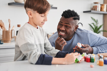 Happy multiracial man smiling toothy and enjoying painting Easter eggs with his caucasian son