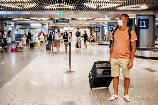 Man in protective medical face mask with backpack and suitcase standing at airport waiting for departure. Male passenger in T-shit waiting for departure gate. Travel during coronavirus virus pandemic