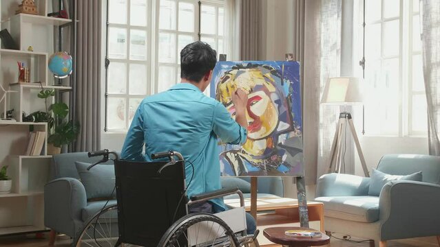 Hind View Of An Asian Artist Man In Wheelchair Holding Paintbrush Mixed Colour And Painting A Girl'S Face On The Canvas
