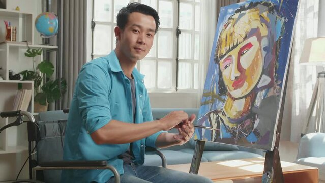 Side View Of An Asian Artist Man In Wheelchair Holding Paintbrush Turn To Wipe The Sweat Before Crossing His Arms And Smiling While Painting A Girl On The Canvas
