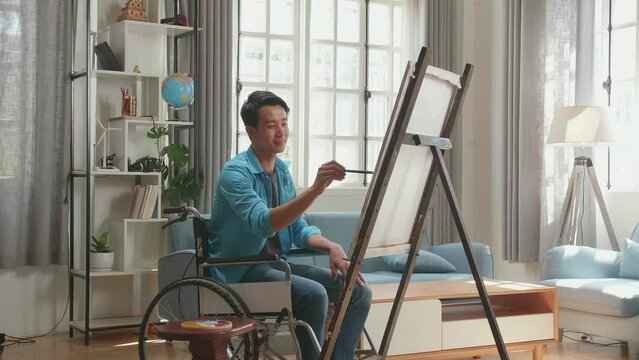 Side View Of An Asian Artist Man In Wheelchair Holding Paintbrush Mixed Colour And Painting On The Canvas
