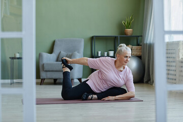 Active senior female practicing yoga asanas on the floor of living-room while sitting on mat and stretching legs and arms