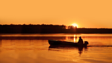 The silhouette of a fisherman in a boat on the river, against the unfocused background of the forest on the opposite bank and the orange dawn