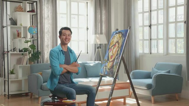 Asian Artist Man Sitting On Round Wooden Chair Without Backrest Holding Paintbrush Mixed Colour, Crossing His Arms, And Smiling After Finish Painting A Girl On The Canvas
